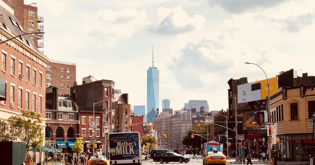 A bustling street scene in Manhattan, New York City, capturing the energy and diversity of urban life. The image showcases a mix of historic brick buildings and modern architecture, with the iconic One World Trade Center standing tall in the background. The street is alive with activity—yellow taxis, delivery trucks, pedestrians, and traffic lights—all under a bright, partly cloudy sky. The scene reflects the dynamic and vibrant atmosphere of one of the world's most famous cities.