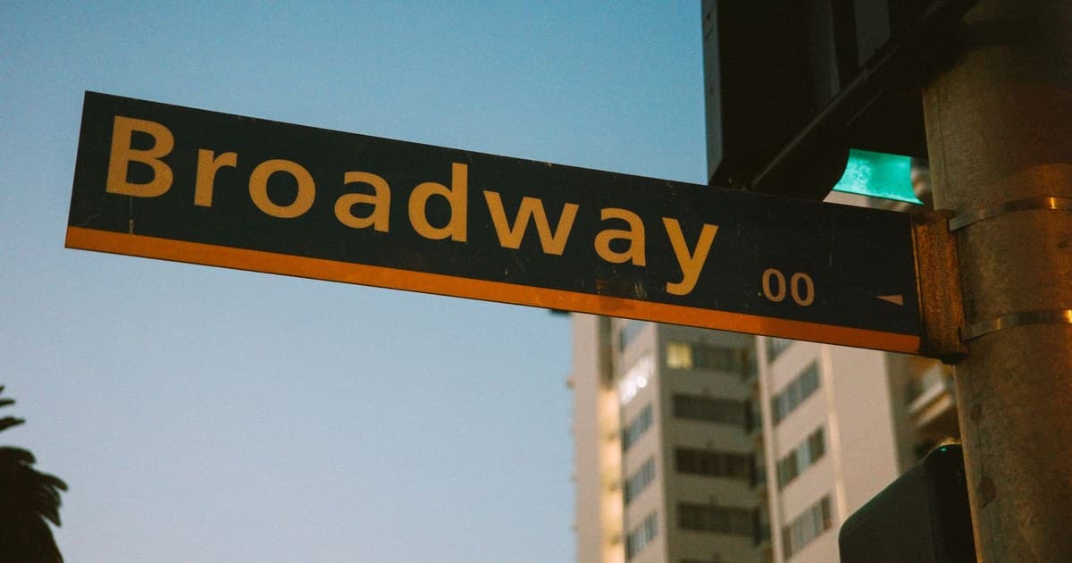 A vibrant street sign depicting Broadway during twilight with a city backdrop.